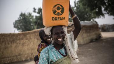 Zwei Frauen lachen; eine von ihnen transportiert einen Oxfam-Wasserkanister auf dem Kopf.
