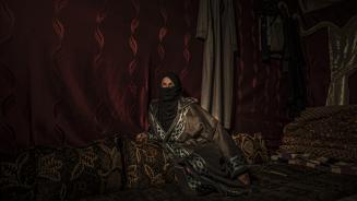 Eine Frau mit langen Gewändern sitzt vor einem roten Vorhang. Durch den Hijab sind nur ihre Augen zu sehen, die interessiert zur Seite schauen.