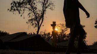 Ein Mensch trägt im Licht der Dämmerung zwei Gegenstände auf seinem Kopf. Der Sonnenuntergang legt sich warm über die Szenerie, ein zweiter Mensch läuft durchs Bild, aber auch dieser Mensch ist nur in seinen Umrissen erkennbar.