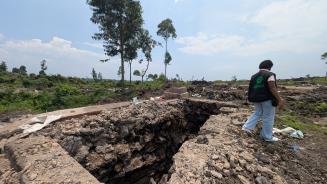 Auf einer flachen, grünen Landschaft sind die Überreste einer zerstörten Latrinenanlage von Oxfam zu sehen. An der Stelle der Latrinen ist nur noch eine tiefe Steingrube übrig. Eine Person, die für Oxfam arbeitet, steht rechts neben der verwüsteten Stelle und sammelt Überreste auf.