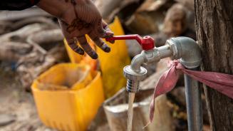 Eine Hand bedient einen Wasserhahn, der an einem Baum festgebunden ist.
