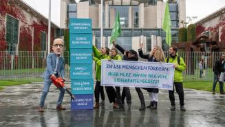Eine Gruppe mit Oxfam-Fahnen vor dem Kanzleramt. Auf ihrem Transparent steht: „210438 Menschen fordern: Herr Merz, Hände weg vom Lieferkettengesetz!“ Eine Person mit Friedrich-Merz-Maske sägt mit einer Kettensäge an einem symbolischen Turm. Der unterste Baustein des Turms ist mit „Lieferkettengesetz“ beschriftet, darüber aufgebaut sind: faire Löhne und sichere Arbeit, Umwelt- und Klimaschutz, Menschenrechte weltweit, globale Gerechtigkeit und zukunftsfähige Wirtschaft.