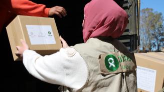 Eine Person mit beiger Oxfam-Weste und rosa Kopftuch lädt Nothilfe-Pakete aus einem Lkw.