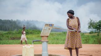 Eine Frau pumpt Wasser in einen Kanister. Hinter transportiert eine andere Frau ein Bündel auf dem Kopf. Im Hintergrund Grün und Nebel