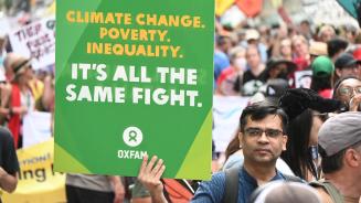 Ein Mann in einer großen Menge Demonstrierender. In der Hand hält er ein Plakat mit der Aufschrift „Climate Change. Poverty. Inequality. It's all the Same Fight.“ (Deutsch: „Klimawandel. Armut. Ungleichheit. Es ist alles derselbe Kampf.")
