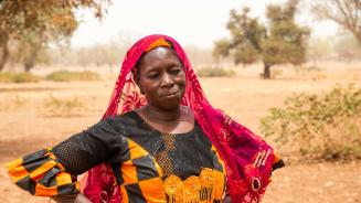 Sanfo Ramata ist Bäuerin und Viehzüchterin. Sie arbeitet für die Confédération Paysanne du Faso, eine Partnerorganisation von Oxfam. Die Frau steht in einem bunten Kleid vor einem Haus. Sie sieht nachdenklich aus.