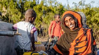 Seadiya Mohamed lächelt in die Kamera, während sie Wasser aus einem laufenden Hahn in ihren Kanister schöpft. Im Hintergrund stehen zwei Kinder.