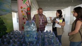 Drei Personen stehen hinter einem großen Stapel Wasserflaschen. Sie halten Klemmbretter in der Hand und sprechen miteinander.