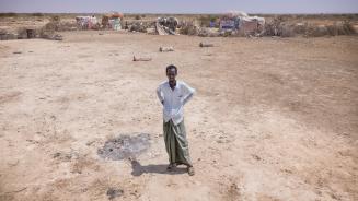 Abdulahi Farah Isse steht, bekleidet mit einem weißen Shirt und einem grauen Wickelrock, auf einem sehr trockenen Boden. Im Hintergrund ist seine Unterkunft zu sehen.