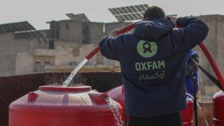 Ein_e Oxfam-Mitarbeiter_in, mit einer blauen Kaputzenjacke mit grünem Oxfam-Logo bekleidet, befüllt mithilfe eines Schlauches einen roten Wassertank mit sauberem Trinkwasser.