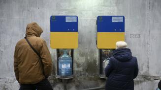 2 Menschen stehen vor zwei Trinkwasserautomaten, aus denen Wasser in ihre Flaschen läuft.