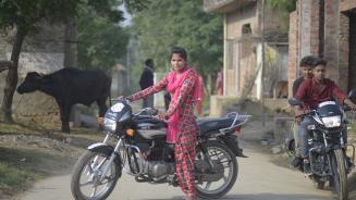 Eine junge indische Frau sitzt auf einem Motorrad; neben ihr zwei Männer auf einem anderen Motorrad