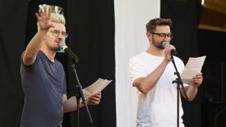 Zwei Männer sprechen in Mikrofone. Sie halten jeweils ein Blatt Papier in der Hand.