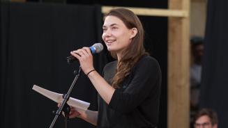 Eine Frau hält lächelnd ein Mikrofon in der einen Hand und Papierblätter in der anderen Hand.