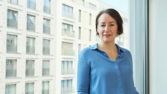 Eine Frau lehnt an einem großen Bürofenster und schaut in die Kamera. Im Hintergrund ein Bürogebäude.