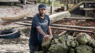 Martin Hau'ato an einem Teil des Schutzwalls, der die Gemeinde Muki auf den Salomonen vor dem steigenden Meeresspiegel schützen soll. Er schaut sehr ernst in die Kamera. Im Hintergrund sieht man das Wasser, das schon fast bis zu den Häusern heran reicht.