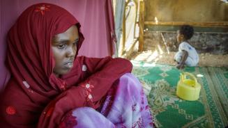 Hafsa Abdikader Ahmed, bekleidet mit einem roten Oberteil und Kopftuch sowie einer lila Hose, sitzt mit herangezogenen Knien auf einem grünen Teppich. Im Hintergrund läuft ein Kind an ihr vorbei. 