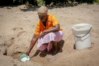 Eine Frau hockt im Sand, vor ihr ein selbst gegrabenes Wasserloch im Boden. Sie schöpft daraus Wasser mit einer kleinen Schüssel. Neben ihr steht ein großer Eimer, der bereits mit Wasser gefüllt ist.