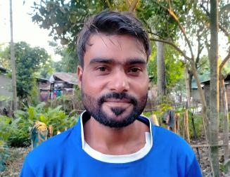 Romeo Lakra auf der Teeplantage in Sonitpur, auf der er lebt und arbeitet. Ein Porträt von Romeo; er trägt ein blaues T-Shirt. Im Hintergrund sind eine Hütte und Pflanzen sowie Bäume zu sehen.