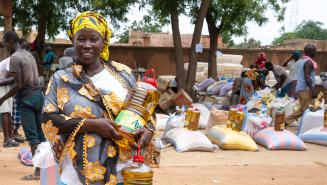 Eine Frau hält zwei große Flaschen Speiseöl. Im Hintergrund liegen vorbereitete Nahrungsmittelpakete bestehend aus Speiseöl und Getreidesäcken.