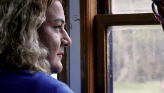 Jennifer schaut aus einem Fenster, sie trägt ein blaues Oberteil und hat schulterlange, wellige Haare.