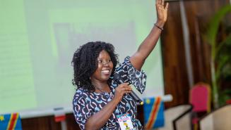 Grâce spricht auf dem Internationalen Afrikanischen Frauenforum (FIFAF) in Kinshasa (Demokratische Republik Kongo). Sie hält ein Mikrofon in der rechten Hand, mit der Linken winkt sie Zuhörenden zu und lächelt sie an. Sie trägt eine blau-gemusterte Tunika.