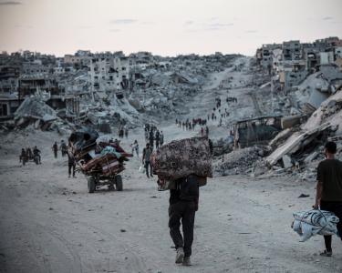 Menschen kehren zurück nach Jabalia in Nord-Gaza. Hier erwartet sie eine katastrophale Zerstörung. Menschen mit Taschen und Beuteln und Karren gehen einen Weg entlang. Rechts und links sind bis zum Horizont Trümmer und zerstörte Häuser zu sehen.