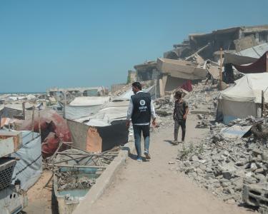Oxfam-Mitarbeiter Osama Qatta zwischen Zelten und Trümmern in Gaza-Stadt. Ein Mann in Oxfam-Weste läuft auf einem schmalen Weg zwischen Trümmern, Steinen, Metallteilen und Zelten. Ein unbekannter Mann kommt ihm entgegen.