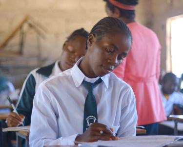 Eine Junge Frau in Schuluniform sitzt an einem Schreibtisch in einem Klassenzimmer und schreibt in ein Heft. Im Hintergrund sind weitere Schüler*innen zu sehen.