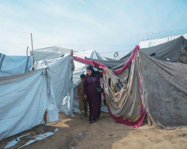 Eine Frau und ein Kind treten hinter einer Decke hervor, die zwischen zwei Holzstelen in einem Geflüchtetencamp gespannt ist. rechts und links erstrecken sich Zelte und weitere gespannte Decken und Tücher.