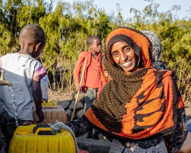 Kommt dank Wasserquelle besser durch den Alltag: Seadiya Mohamed Seadiya Mohamed lächelt in die Kamera, während sie Wasser aus einem laufenden Hahn in ihren Kanister schöpft. Im Hintergrund stehen zwei Kinder.