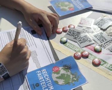 Eine Person füllt eine Unterschriftenliste aus, die neben einem Flyer „Reichtum fairteilen“, Oxfam-Buttons und „Tax-the-rich“-Tattoos liegt