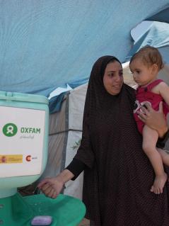 Duaa Abu Sabha wäscht sich die Hände an einer Handwaschstation, die in einem Camp für Geflüchtete steht. Auf dem Arm trägt sie ein Kleinkind.