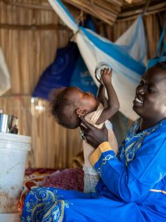 Ayul Ajak sitzt in einer provisorischen Unterkunft und spielt mit einem Kleinkind. Im Hintergrund steht ein Eimer mit der Aufschrift „Oxfam“.