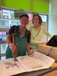 Ein Mann und eine Frau stehen lächelnd hinter der Kasse eines Oxfam Shops. Der Mann hält eine Vase in der Hand. 