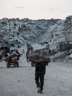 Menschen kehren zurück nach Jabalia in Nord-Gaza. Hier erwartet sie eine katastrophale Zerstörung. Menschen mit Taschen und Beuteln und Karren gehen einen Weg entlang. Rechts und links sind bis zum Horizont Trümmer und zerstörte Häuser zu sehen.