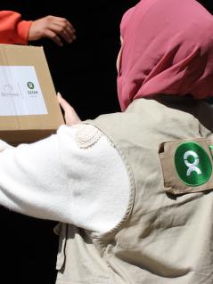 Eine Person mit beiger Oxfam-Weste und rosa Kopftuch lädt Nothilfe-Pakete aus einem Lkw.