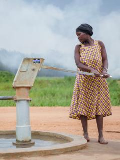 Eine Frau pumpt Wasser in einen Kanister. Hinter transportiert eine andere Frau ein Bündel auf dem Kopf. Im Hintergrund Grün und Nebel