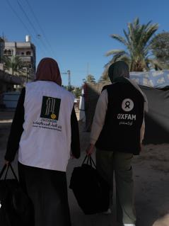 Eine Mitarbeiterin unserer Partnerorganisation Juzoor for Health and Social Development ist gemeinsam mit einer Oxfam-Kollegin in Gaza im Einsatz. Zwei Frauen mit großen Taschen in Rückenansicht. Eine trägt eine Weste der Organisation Juzoor, die andere eine Weste von Oxfam.