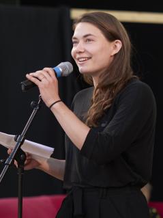 Eine Frau hält lächelnd ein Mikrofon in der einen Hand und Papierblätter in der anderen Hand.