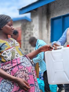 Muhawe Myriam Charmante erhält einen Wasserkanister von einem Oxfam-Mitarbeiter. Sie lebt in einem Camp für Geflüchtete in Sake, Demokratische Republik Kongo. Ein Mann in Oxfam-Weste reicht einer schwangeren Frau einen weißen Wasserkanister.