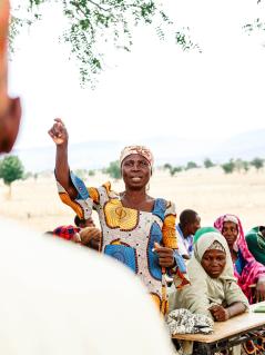 Teilnehmer*innen eines Projekts zu Geschlechtergerechtigkeit und politischer sowie ökonomischer Teilhabe im nigerianischen Bundesstaat Adamawa. Ein Mann steht draußen mit dem Rücken zur Kamera. Ihm gegenüber steht eine Frau, die gerade einen Arm hebt und etwas sagt. Hinter ihr sitzen mehrere Leute an Schreibtischen.