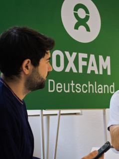 Die Oxfam-Mitarbeitenden Caspar Altewischer und Jörn Kalinski (von links) im Gespräch miteinander: Sie tauschen sich anllässlich von 30 Jahren Oxfam Deutschland dazu aus, was der Oxfam Deutschland e.V. seit 1995 bewirkt und bewegt hat. Zwei Männer im Gespräch. Sie sitzen vor einem Schild mit der Aufschrift „Oxfam Deutschland.“.