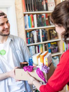 Eine Frau übergibt einem Mann einen Karton mit Büchern, Halstüchern und anderen Dingen.