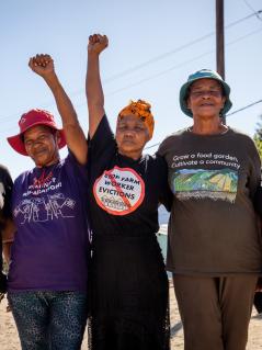 Gruppenbild von WoFP-Aktivistinnen Gruppenbild von Women on Farms Project Aktivistinnen. Sie strecken ihre Fäuste in die Luft. In der Mitte ist Bettie Fortuin zu sehen, sie trägt ein gelbes, geknotetes Kopftuch und ein schwarzes T-Shirt mit der Aufschrift "Stop Farm Worker Evictions"