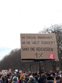 viele Menschen mit Demoschildern bewegen sich auf den Großen Stern in Berlin zu. Auf einem Schild steht "Die einzige Minderheit, die die Welt ruiniert, sind die reichsten 1 %"