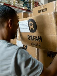 Ein Mann hält einen Karton mit Oxfam-Logo. Im Hintergrund sind Regale mit Nahrungsmitteln zu sehen.