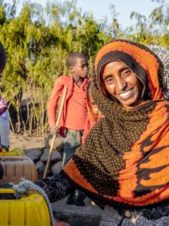 Seadiya Mohamed lächelt in die Kamera, während sie Wasser aus einem laufenden Hahn in ihren Kanister schöpft. Im Hintergrund stehen zwei Kinder.