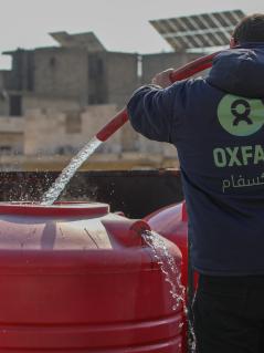 Ein_e Oxfam-Mitarbeiter_in, mit einer blauen Kaputzenjacke mit grünem Oxfam-Logo bekleidet, befüllt mithilfe eines Schlauches einen roten Wassertank mit sauberem Trinkwasser.