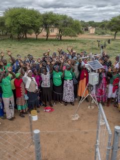 Eine Gruppe von Menschen steht bei einer Wetterstation in einer ländlichen Region in Simbabwe. Sie strecken freudig ihre Arme in die Luft.